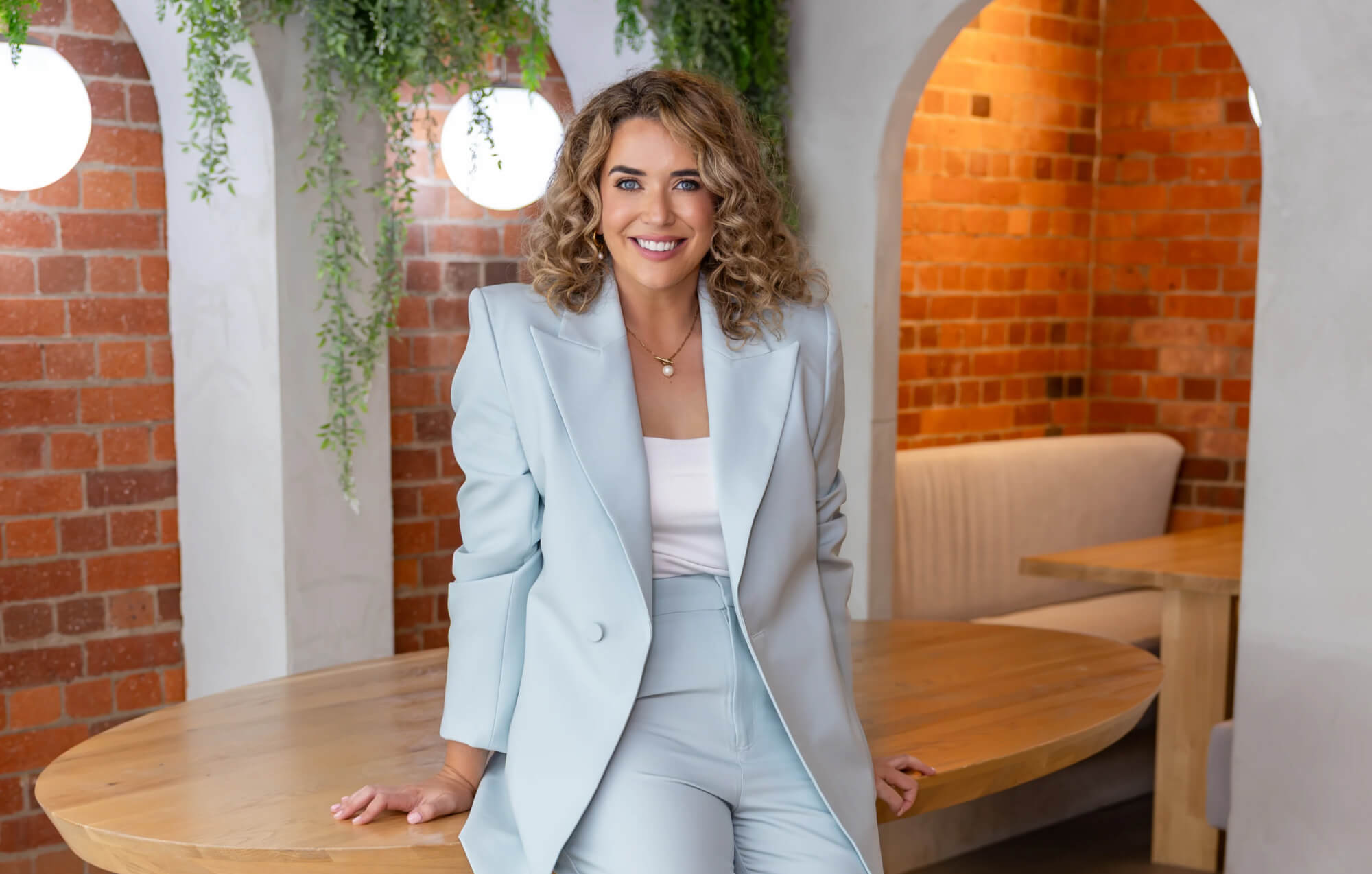 Caitlin in a light blue suit standing in a modern cafe with brick walls and wooden tables.