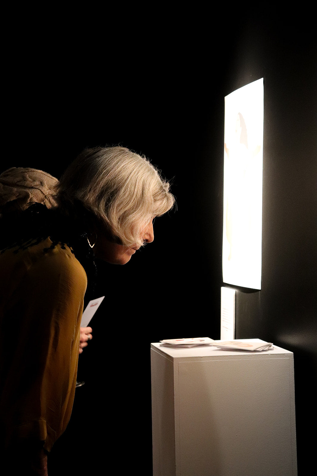 A woman with short gray hair leans in toward a glowing vertical light panel mounted above a white pedestal in a dark room.