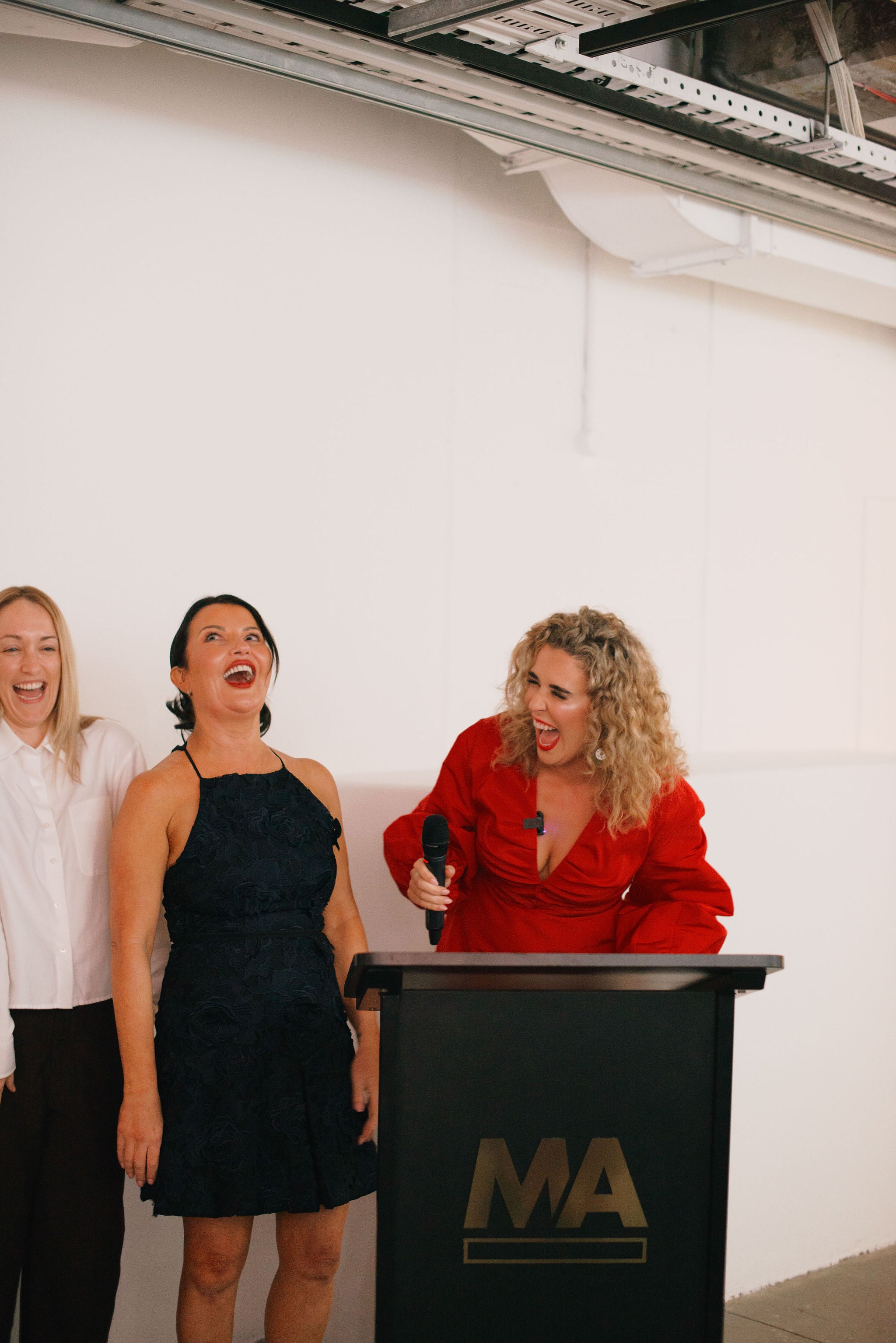 Caitlin in a red top is speaking behind a black podium featuring a gold 'MA' logo. Two other women are standing to the left.